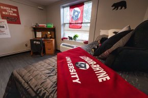 Model dorm room with bed and red LR blanket in close foreground, window and desk further back