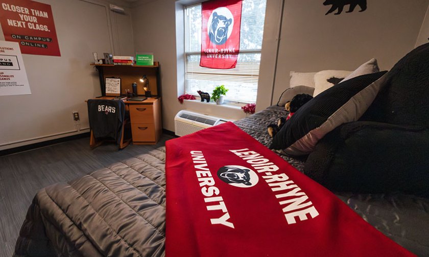 Model dorm room with bed and red LR blanket in close foreground, window and desk further back