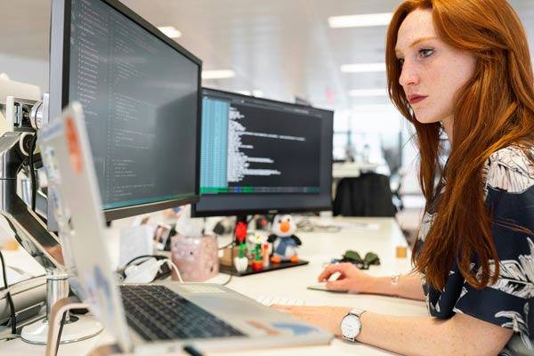 Woman working at an office desk reviewing computer code on two monitors