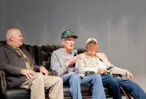 Three men sit on an indoor stage