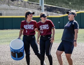 Two softball players laugh on the field with Coach Shena Hollar