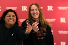 In front of red backdrop with LR logo visible, two female students hold out Bears Say Thanks stickers to foreground 