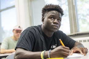 A student with pencil in hand looking past the camera toward the front of a classroom