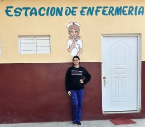 Alyssa Bailey stands by a clinic door under a mural of a nurse and the words "estacion de enfermeria" above