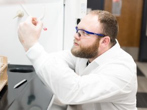 Noah Jenkins wears a lab coat and safety glasses in an indoor lab