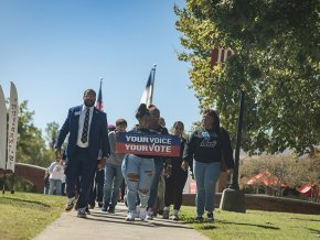 A group of LR students, faculty and staff walk down a sidewalk away from Shaw Plaza, holding a sign in front that reads "Your Voice, Your Vote"