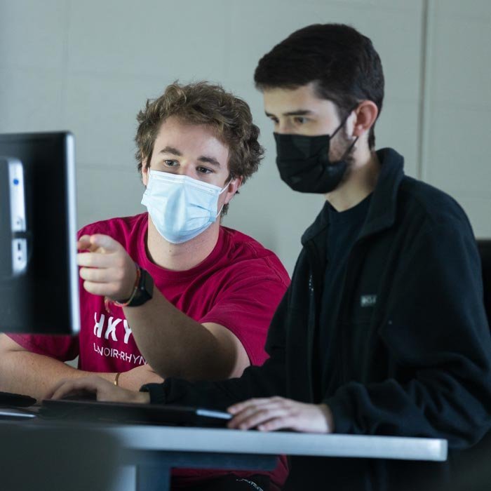 Two masked male students working on computer assignment in classroom